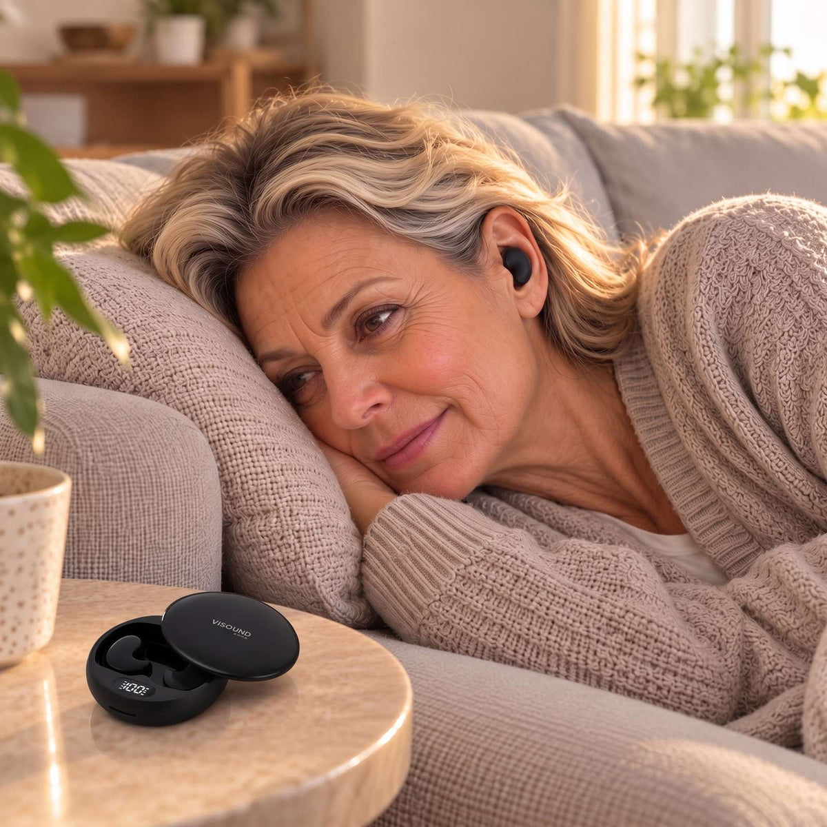 Woman around 60 lying on the couch with ViSound Drift flat in her ear, glossy case on the side table
